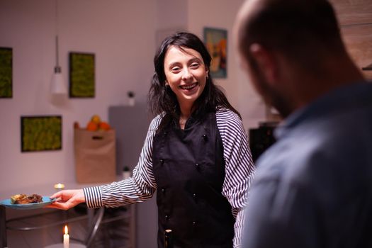 Wife smiling at husband wearing apron while looking at husband after cooking tasty dinner. Woman cooking festive food for her and husband to celebrate relationship and romance, happiness, positive, love, emotion, marriage.