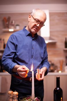 Senior man lighting candle with matches in kitchen for romantic dinner with wife. Elderly old husband preparing festive meal with healty food for anniversary celebration, sitting near the table.