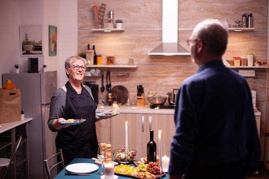 Relationship celebration in kitchen with senior cheerful couple. Elderly old couple talking, sitting at the table in kitchen, enjoying the meal, celebrating their anniversary with healty food.