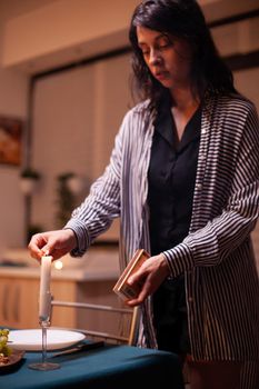 Young lady lighting the candles on kitchen table. Caucasian happy wife preparing festive meal with healthy food for anniversary celebration, sitting near the table in kitchen.