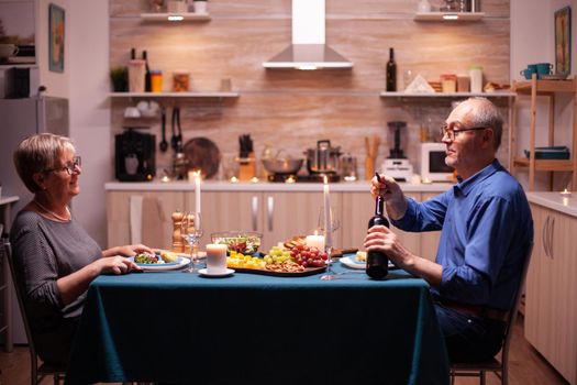 Senior man opening wine bottle in kitchen while celebrating relationship with wife enjoying tasty meal. Elderly old couple talking, sitting at the table in kitchen,