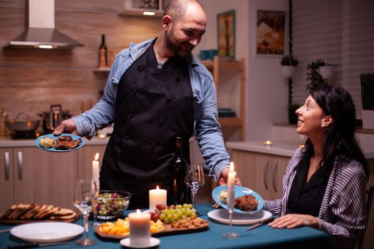 Happy wife smiling at husband during romantic celebrating relationship. Man preparing festive dinner with healthy food, cooking for his woman a romantic dinner,