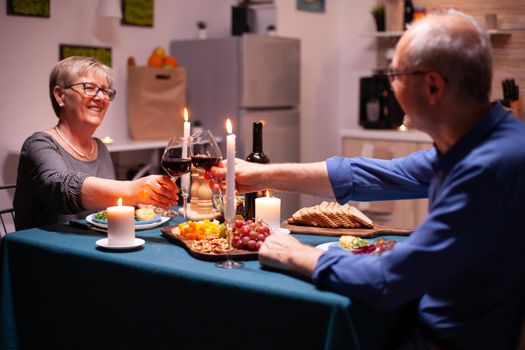 Senior couple celebrating relationship in evening toasting glasses with red wine. Elderly man and woman sitting at the table in kitchen, talking, enjoying the meal,