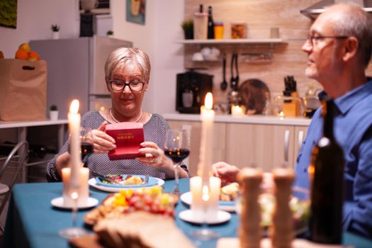 Retired woman holding gift box from husband during relationship aniversary dinner. Happy cheerful elderly couple dining together at home, enjoying the meal, celebrating their marriage , surprise holiday