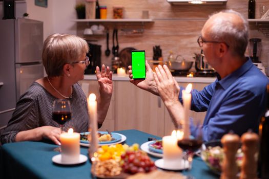 Aged man holding phone with green screen and wife is waving at it. Aged people looking at mockup template chroma key isolated smart phone display using techology internet sitting at the table