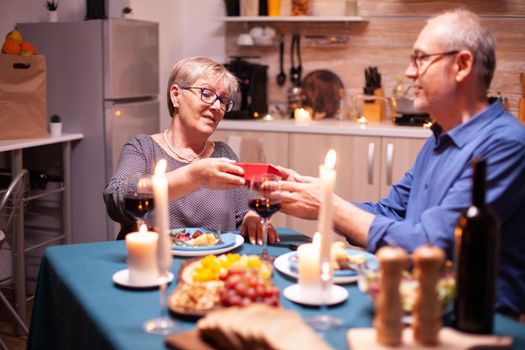 Elderly husband giving wife gift box during dinner. Happy cheerful elderly couple dining together at home, enjoying the meal, celebrating their anniversary, surprise holiday