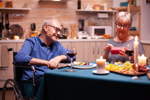 Grandmother looking at gift from disabled husband in wheelchair. Happy cheerful elderly couple dining together at home, enjoying the meal, celebrating their marriage.