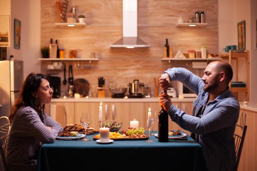 Husband opens wine bottle while celebrating relationship with wife in kitchen. Happy couple talking, sitting at table in kitchen, enjoying the meal, celebrating their anniversary at home.