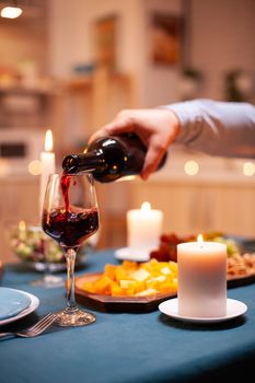 Close up of guy pouring wine in glass during romantic dinner with wife in dining room. Young man pouring red wine in wife glass. Romantic caucasian happy couple sitting at the table celebrating .