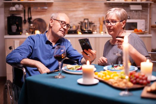 Man in wheelchair holding phone while having dinner with wife in kitchen. Scrolling and showing photos. Imobilized handicapped senior husband scolling on phone enjoying the festive meal.