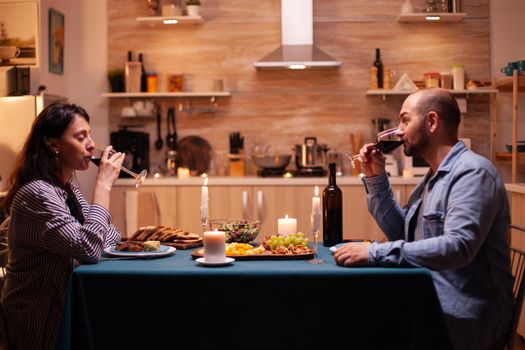 Wife with husband drinking red wine enjoying romantic dinner. Relax happy people ,sitting at table in kitchen, enjoying the meal, celebrating anniversary in the dining room