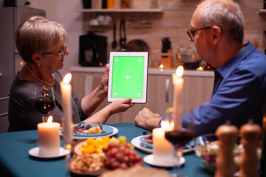 Elderly husband and wife looking at tablet pc with green mockup. Aged people looking at green screen template chroma key display sitting at the table in kitchen during dinner.