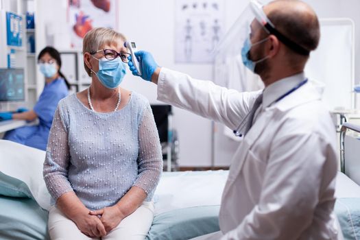 Doctor wearing protective mask against coroanvirus checking patient temperature using infrared thermometer. Medical examination for infections, disease and diagnosis.