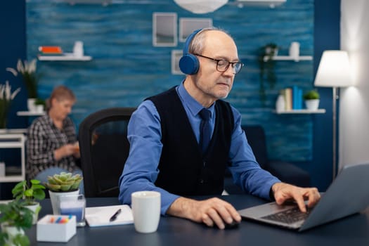 Senior businessman listening music wearing headphones working on laptop. Elderly man entrepreneur in home workplace using portable computer sitting at desk while wife is reading a book sitting on sofa.