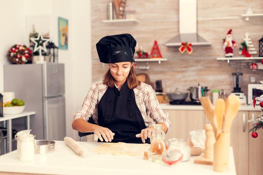 Sweet child making dessert on christmas day wearing apron. Cheerful happy cute girl while prepearing delicious cookies for christmas celebration in kitchen with christmas tree in the background.