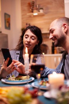 Wife showing husband photos on smartphone during festive relationship anniversary. Adults sitting at the table in the kitchen browsing, searching, using smartphones, internet, celebrating