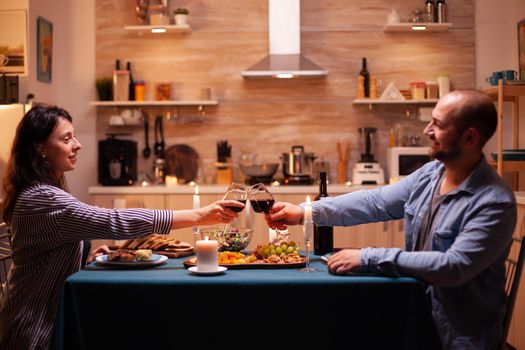 Positive husband and wife holding wine glasses in kitchen in the evening. Relax happy people clinking, sitting at table in kitchen, enjoying the meal, celebrating anniversary in the dining room.