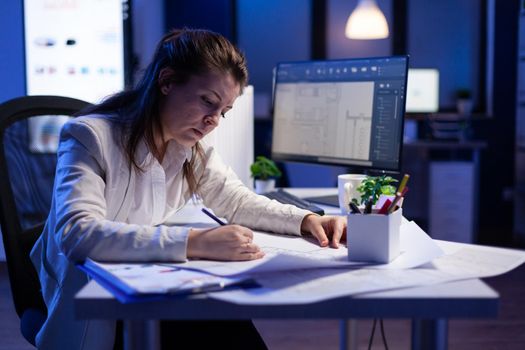 Overworked woman architect checking and matching blueprints sitting at office desk in front of computer. Perfectionist designer using arhitecture blueprints of buildings creating industrial prototype
