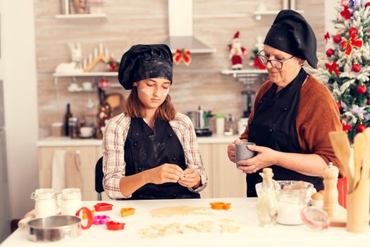 Grandchild learning to make dough on christmas day from grandmother with apron. Happy cheerful joyfull teenage girl helping senior woman preparing sweet cookies to celebrate winter holidays.