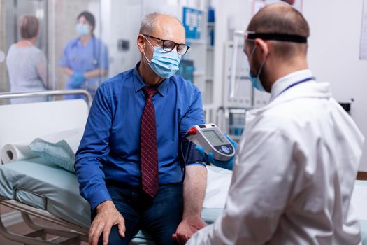 Doctor wearing protective mask agasint coronavirus checking blood pressure of senior patient in hospital examination room. Medical examination for infections, disease and diagnosis.