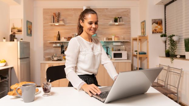 Freelance businesswoman sitting at desk in home working on laptop. Concentrated entrepreneur in home kitchen using notebook during late hours in the evening.