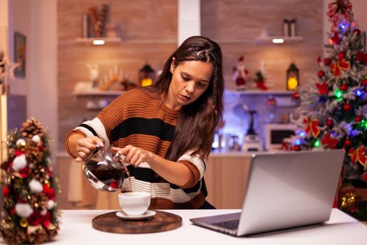 Caucasian woman pouring cup of tea from kettle while talking to friends on video call app. Young adult using laptop for christmas eve in holiday decorated kitchen with ornaments