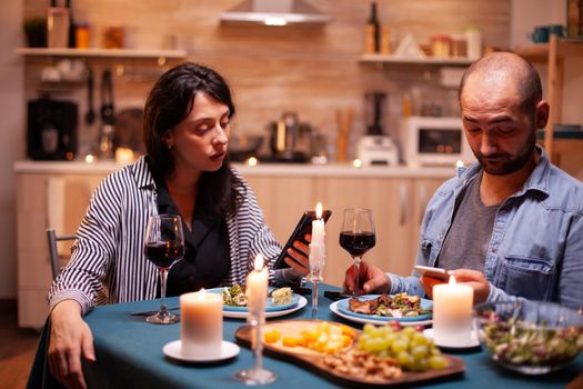 Happy couple watching video on smartphone in kitchen during anniversary. Adults sitting at the table in the kitchen browsing, searching, using smartphones, internet, celebrating anniversary.