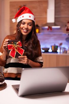 Caucasian woman opening present on video call conference technology with friends in festive home. Cheerful young person receiving surprise gift for christmas eve seasonal celebration