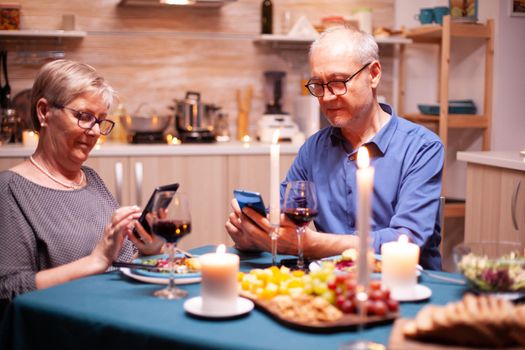 Browsing on smartphone in during their relationship anniversary in kitchen with candles on table. Sitting at the table in the kitchen, browsing, searching, using phone, internet, in the dining room.