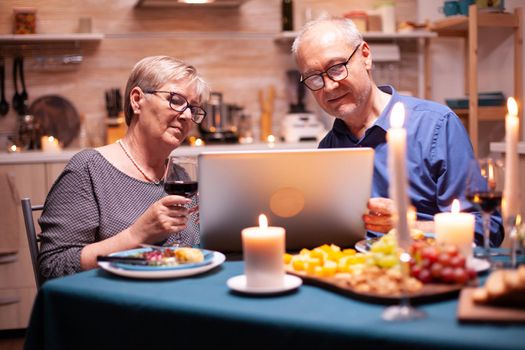 Senior woman holding wine glass and using laptop in kitchen with husband and festive food on table. Elderly people sitting at the table browsing, searching, using laptop, technology, internet, celebrating their anniversary in the dining room.