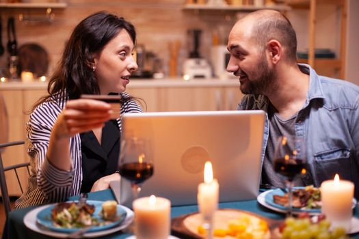 Couple looking at each other while doing online shopping during romantic dinner. Adults sitting at the table, searching, browsing, surfing, using technology card payment, internet