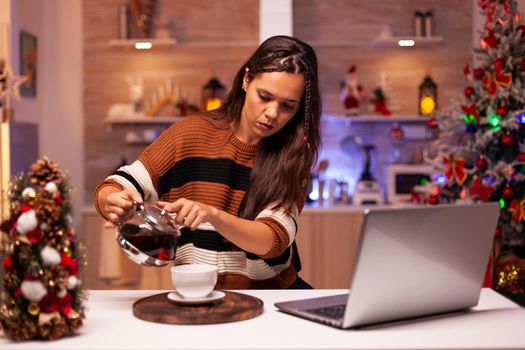 Modern woman filling mug of tea from kettle on video call connection talking to friends. Cheerful young person using laptop for wishing merry christmas in seasonal decorated kitchen