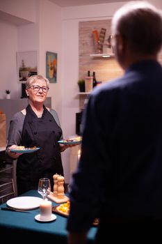 Senior couple having a conversation in kitchen. Elderly aged woman wearing apron. Elderly old couple talking, sitting at the table in kitchen, enjoying the meal, celebrating their anniversary