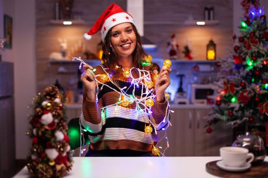 Happy young woman trying to decorate tree with lights on string for christmas holiday in festive kitchen at home. Caucasian smiling person knotted in bright tangled garland wire bulbs