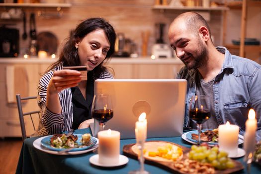 Couple doing shopping online at dinner on laptop. Adults sitting at the table, searching, browsing, surfing, using technology card payment, internet