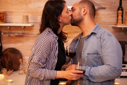 Happy couple kissing in kitchen during anniversary of their relationship. Adult couple at home, drinking red wine, talking, smiling, enjoying the meal in dining room.