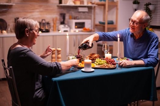 Senior man pouring wine to wife while celebrating relationship anniversary in kitchen. Romantic couple sitting at the table in dining room , talking, enjoying the meal.