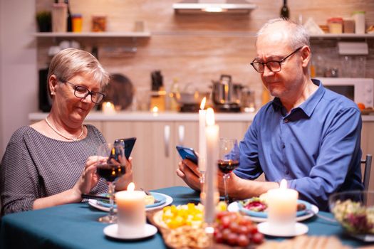 Elderly man scrolling on phone for memories to show her wife during romantic date. Sitting at the table in the kitchen, browsing, searching, using phone, internet, celebrating their anniversary.