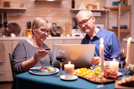 Elderly woman holding credit card while using laptop with husband for shopping. Old people sitting at the table, browsing, using the technology, internet, celebrating their anniversary.