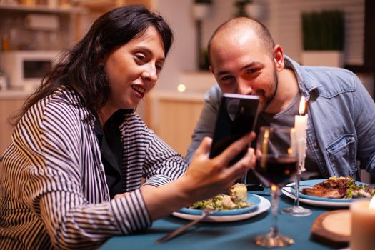 Happy couple watching video on smartphone in kitchen during anniversary. Adults sitting at the table in the kitchen browsing, searching, using smartphones, internet, celebrating anniversary.