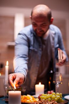 Husband lighting candle on kitchen table a for festive dinner with wife. Man preparing festive meal with healthy food for anniversary celebration, romantic date.