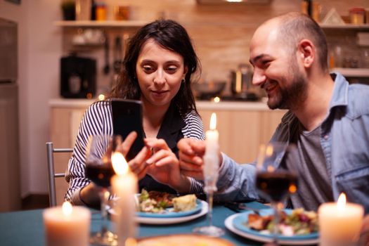 Couple using phone during romantic dinner in kitchen and candles burning on table. Adults sitting at the table in the kitchen browsing, searching, using smartphones, internet, celebrating anniversary.