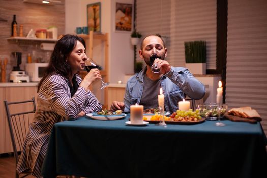 Couple drinking red wine having relationship celebration. Happy couple talking, sitting at table in dining room, enjoying the meal, celebrating their anniversary at home having romantic time.