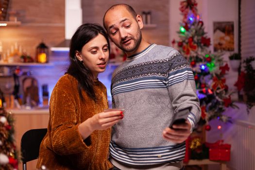 Cheerful family doing online shopping xmas gift using credit card for payment on smartphone celebrating christmas holiday. Happy couple enjoying winter season spending christmastime together