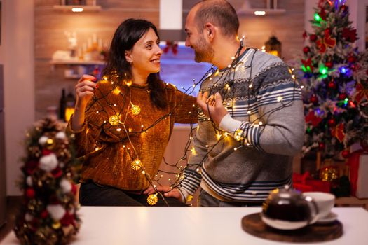 Amused couple having fun with christmas tree light decorating kitchen enjoying spending wintertime together. Happy family celebrating santa-claus festive season. Christmastime tradition