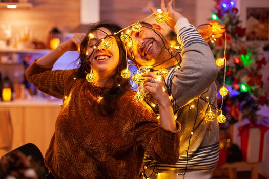 Funny couple caughting in christmas tree light during christmastime preparing decoration for kitchen. Happy family enjoying spending winter holiday together. Santa-claus festive tradition