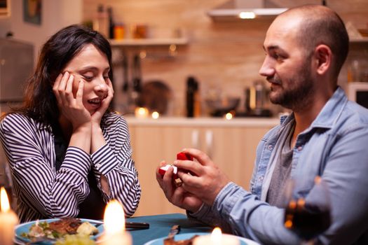 Man celebrating relationship and asking girlfriend to marry him while having dinner. Man asking his girlfriend to marry in the kitchen during romantic dinner. Happy caucasian woman smiling being speechless