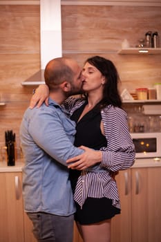 Husband kissing wife while dancing in kitchen. Happy in love couple dining together at home, enjoying the meal, smiling, having fun, celebrating their anniversary.