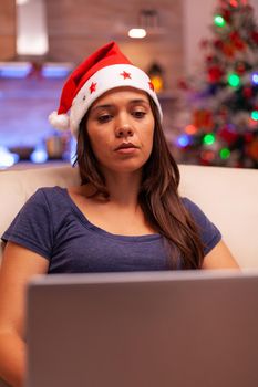 Girl looking at laptop screen reading business email during christmas holiday sitting comfortable on sofa in xmas decorated kitchen. Woman adult browsing on social media. Winter season
