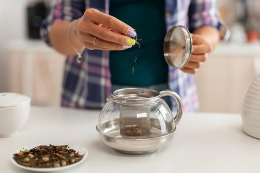 Close up of pouring aromatic herbs in teapot to make tea in morning for breakfast. Preparing tea in the morning, in a modern kitchen sitting near the table. Putting with hands, healthy herbal in pot.